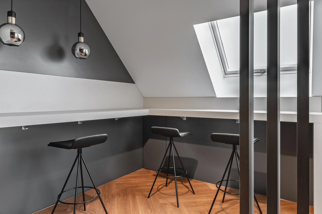 Thekenbereich unter der Dachschr&auml;ge mit weisser Arbeitsplatte, drei schwarzen Hockern, schr&auml;ger Fenster&ouml;ffnung, zwei Pendelleuchten und Holzfussboden
