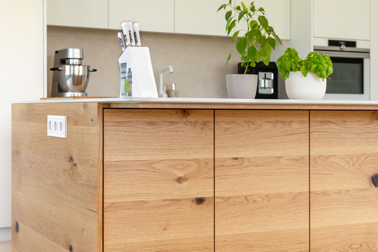Detailaufnahme einer K&uuml;cheninsel mit furnierter Holzoberfl&auml;che, weisser Arbeitsplatte, Steckdosen, Messerblock, Kr&auml;utertopf und Kaffeemaschine im Hintergrund