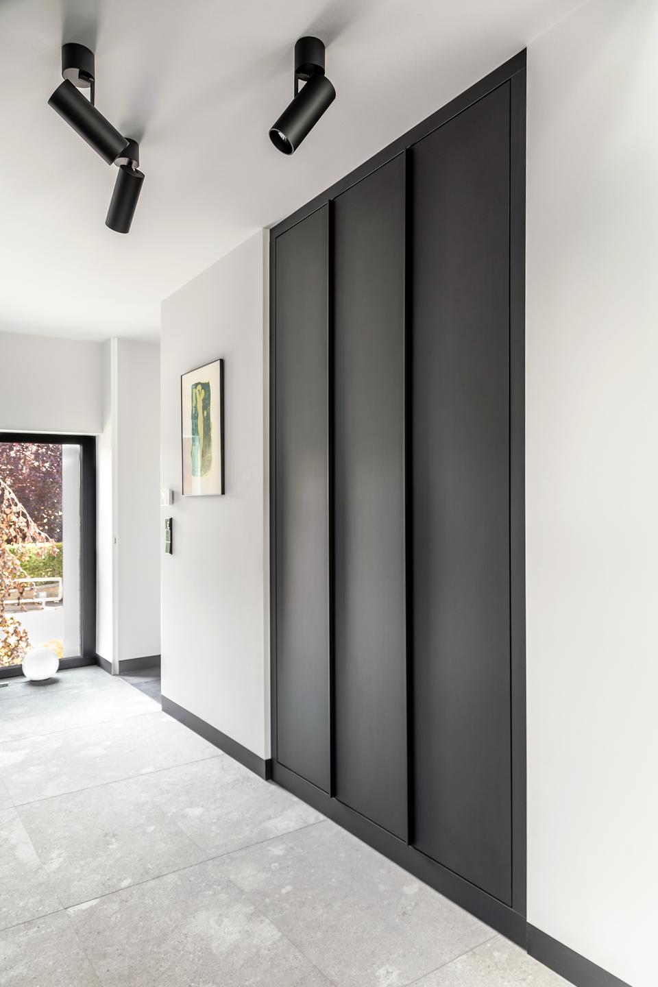 Schwarzer Wandschrank mit vertieften Fronten in einem Eingangsbereich Eingangsbereich mit grossformatigem schwarzen Wandschrank, vertieften Fronten, Deckenstrahlern und grauem Steinboden